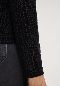 Maglia nera a maniche lunghe con un motivo a coccodrillo testurizzato, realizzata in un materiale morbido. Presenta una finitura leggermente trasparente al polsino.