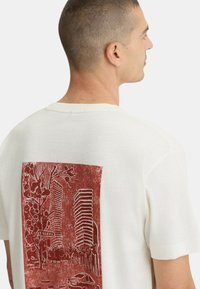 T-shirt bianco in cotone con una stampa grafica rossa sul retro, che rappresenta edifici e alberi di città, con dettagli di linea chiari.