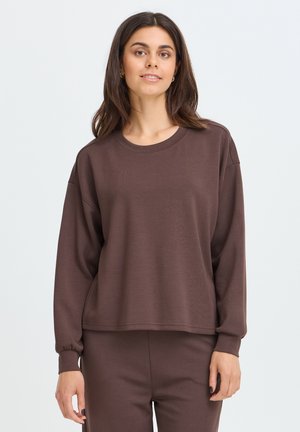 Kvinde med mørkt hår iført en løs mørkebrun langærmet sweatshirt og matchende bukser mod en ensfarvet lys baggrund.