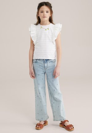 Bluză albă din bumbac cu mâneci volănaș, asociată cu blugi din denim brodați de culoare albastru deschis și saboți maronii cu platformă din lemn.