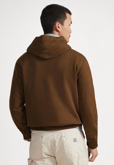 Άνδρας με πλάτη, φοράει καφέ φούτερ με κουκούλα πάνω από γαλάζιο πουκάμισο και μπεζ παντελόνι με πίσω τσέπες.