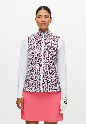 Donna che indossa una maglia bianca a maniche lunghe con un gilet leopardato rosa e nero, gonna rosa e guanto da golf bianco sulla mano destra.