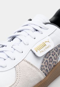 Baskets blanches avec des accents en cuir suédé et imprimé léopard, ainsi que des marquages dorés. Elles possèdent des lacets blancs, une texture lisse et une semelle en caoutchouc marron.