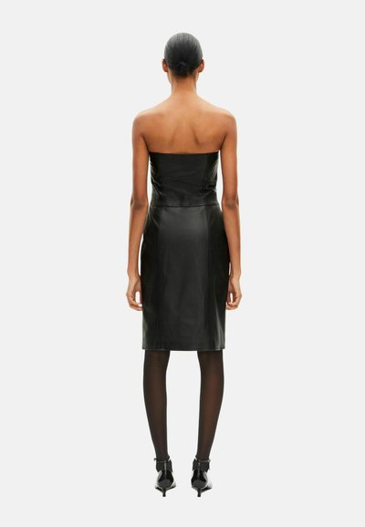 Robe noire en cuir sans bretelles avec une silhouette ajustée, un ourlet arrivant au genou et une texture lisse. Portée avec des collants noirs transparents et des talons pointus.