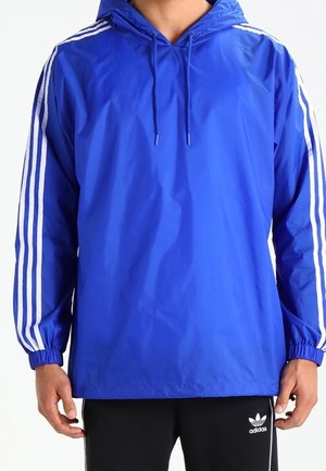 Man die een blauwe windjack met capuchon draagt met witte strepen op de mouwen en zwarte Adidas-broek met logo op de linker dij.