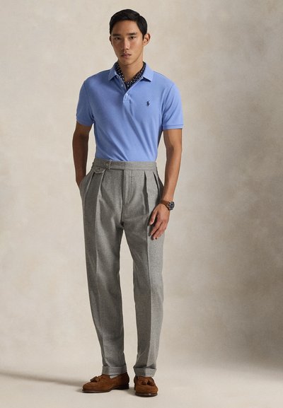 Polo azul claro, calças cinza plissadas, mocassins castanhos. Detalhes notáveis: gola com bolas e logótipo subtil na camisola.