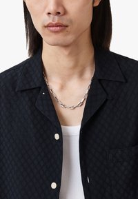 Chemise noire texturée avec un motif de feuilles, dotée d'un col, d'une fermeture à bouton, et d'un collier en chaîne argenté sur un débardeur blanc à côtes.