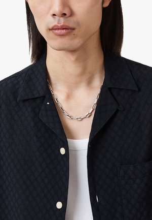 Black textured shirt with a leaf pattern, featuring a collar, button closure, and a silver chain necklace over a white ribbed tank top.