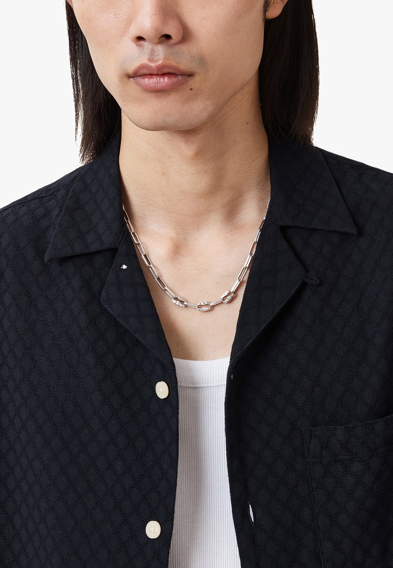 Chemise noire texturée avec un motif de feuilles, dotée d'un col, d'une fermeture à bouton, et d'un collier en chaîne argenté sur un débardeur blanc à côtes.