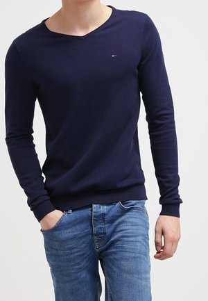 Mann trägt einen marineblauen V-Ausschnitt-Pullover mit langen Ärmeln und hellblaue Jeans, steht mit einer Hand in der Tasche.