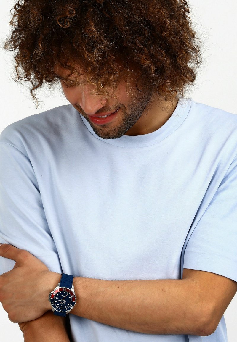 Camicia azzurra a maniche corte indossata con un orologio dotato di un cinturino in gomma blu scuro e un quadrante rotondo con accenti rossi e argentati.