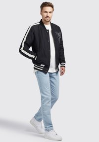 Zwarte bomberjack met witte strepen, geborduurd ontwerp, met drukknopen aan de voorkant, gecombineerd met lichtblauwe jeans en witte sneakers.