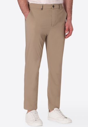 Uomo che indossa pantaloni beige slim-fit con tasche laterali, chiusura con bottone e zip, abbinati a sneakers bianche e camicia bianca.