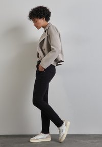 Béžová zkrácená bunda s dvojitými knoflíky, černé slim-fit džíny a bílé tenisky s béžovými detaily. Modelka pózuje bokem proti šedé zdi.