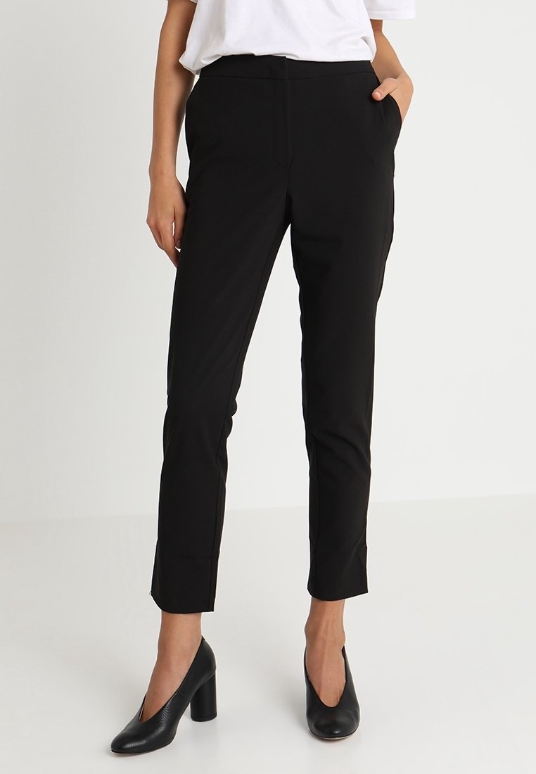 Femme portant un pantalon noir slim, un haut blanc et des chaussures noires à talons hauts, debout avec une main dans la poche sur fond blanc.