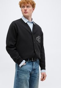 Νέος άνδρας με σγουρά μαλλιά, που φοράει μαύρη ζακέτα με το λογότυπο "cK" πάνω από μπλε ριγέ πουκάμισο και μπλε τζιν, στέκεται μπροστά σε απλό φόντο.