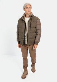 Gepolsterte Jacke aus einer Mischung von olivfarbenem Stoff und braunen Ärmeln, mit einem Reißverschluss vorne und einem kragen aus hellbraunem Fleece. Braune Hose und Stiefel.