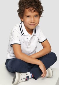 Witte poloshirt met marineblauwe accenten en een strikjespatroon, marineblauwe broek en witte sneakers met blauw/rode gestreepte sokken. Jongen die in kleermakerszit zit.