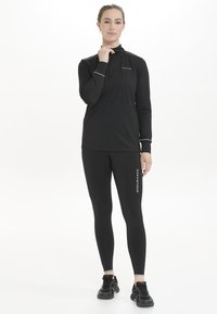 Sort langærmet zip-up trøje med refleksaccentuer, parret med sorte leggings med "ENDURANCE" trykt i hvid. Atletisk fodtøj fuldender looket.