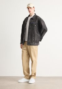 Giacca di jeans nera con tasche frontali, t-shirt grigia, pantaloni beige e sneakers bianche. Il cappellino è di colore crema con un logo. Design minimalista.