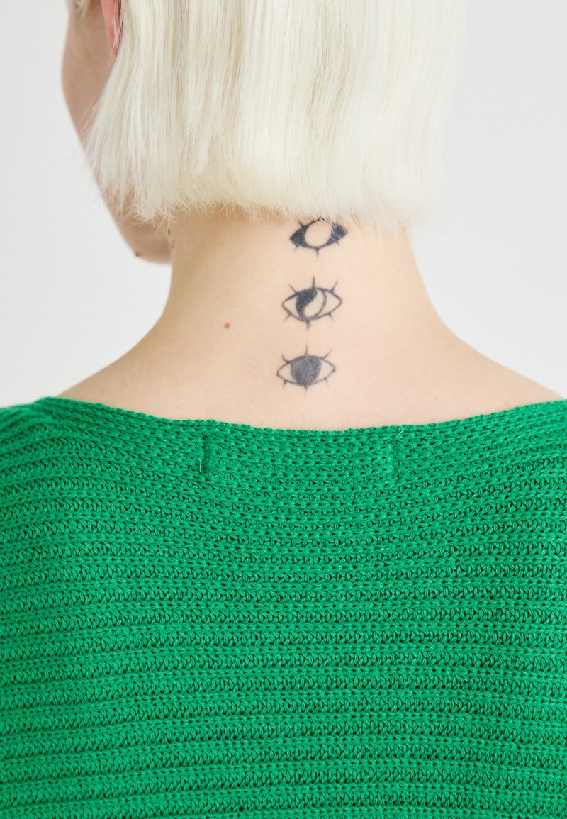 Grüner Strickpullover mit einem strukturierten Muster, getragen von einer Person, die drei schwarze Augentattoos am Nacken zeigt.