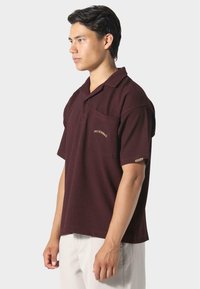 Polo shirt bordeaux a maniche corte in un tessuto strutturato, con taschino sul petto e testo ricamato. Vestibilità comoda con colletto.