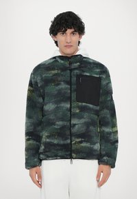 Fleece jacket in dark green with abstract patterns. Features a black zipped pocket, high collar, and a relaxed fit. Soft texture.