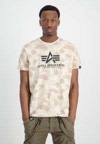 Beige camouflage T-shirt med korta ärmar, har svart "Alpha Industries"-logga på bröstet, slätt tyg, rund halsringning.