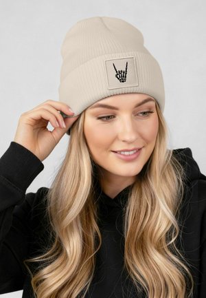Cream ribbed beanie with a square patch featuring a black skeleton hand graphic. Made of soft material, suitable for warm wear.