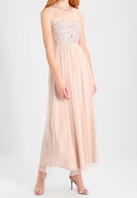 Lång, naken kvällsklänning med en pärlad blom-bodice, genomskinliga axelband och flödande tylskjol. Har en subtil lyster och en elegant silhuett.