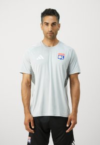 Lys grå sportsjakke med korte ermer, med Olympique Lyonnais-logoet og svarte detaljer. Laget av fukttransporterende stoff.