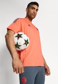 Koral fotsportskjorte med mørke skulderstriper, kombinert med grå bukser som har en rød FC Bayern München-logo, og holder en fotball med stjernesymboler.