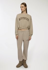 Felpa cropped beige con "KENTUCKY" in marrone, abbinata a pantaloni della tuta beige coordinati con un piccolo logo. Stivali in suede marroni completano l'outfit.