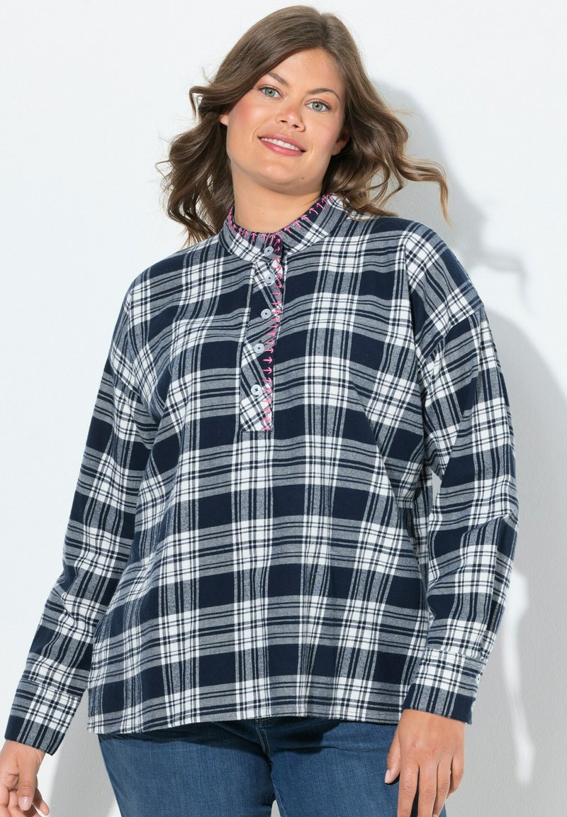 Donna che indossa una camicia a quadri blu e bianca con dettagli in cucitura rosa, abbinata a jeans blu, sorridente con capelli ondulati su sfondo bianco.