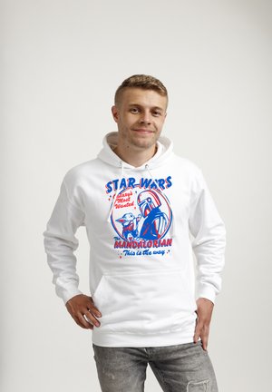 Ung mand iført hvid hættetrøje med rød og blå Star Wars Mandalorian grafik og slogan, stående mod en ensartet lys baggrund.