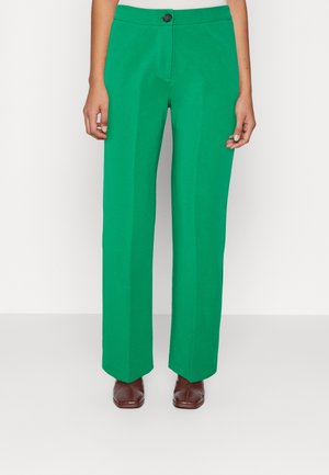 Groene broek met hoge taille en wijde pijpen met een vouw aan de voorkant, zwarte knoopsluiting, gedragen met bruine schoenen met vierkante neus en een wit ingestopt topje.