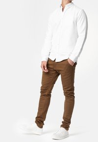 Hvid langærmet skjorte med knappetalje og brystlomme, parret med slanke brune bukser og hvide sneakers, der viser en moderne afslappet stil.