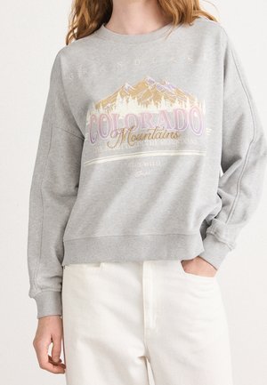 Person iført en grå sweatshirt med et Colorado Mountains-motiv og hvide bukser, hånd i lommen, mod en ensfarvet baggrund.