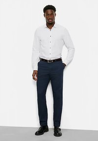White long-sleeve shirt with black buttons, paired with navy trousers and black shoes; smooth fabric, tailored fit, and no visible patterns.