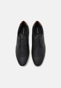 Zapatos de cuero perforado negros con detalles en marrón, punta redonda, diseño de cordones y suela de goma blanca. El interior presenta la etiqueta de la marca.
