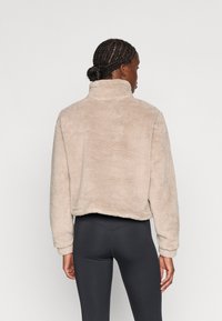 Veste en fausse fourrure beige avec un col haut, une coupe cropped et des manches longues, assortie à des leggings noirs. Texture douce et coupe décontractée.
