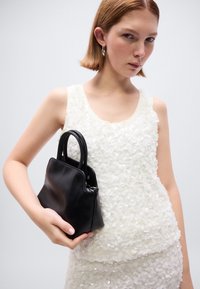 Μαύρη δερμάτινη τσάντα χειρός με στρογγυλεμένες λαβές, λεία υφή και γωνιακό σχήμα. Το μοντέλο φοράει λευκό τοπ διακοσμημένο με κυκλικά, λαμπερά πέταλα.