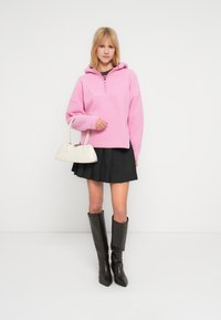 Νεαρή γυναίκα με ροζ φλις φούτερ με κουκούλα, μαύρη πλισέ φούστα, μαύρες μπότες μέχρι το γόνατο, κρατώντας μια μικρή λευκή τσάντα, στέκεται μπροστά σε λευκό φόντο.