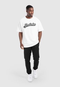 Camiseta de algodón blanca con el logo negro "Smilodox"; combinada con pantalones negros y zapatillas blancas; diseño simple y de corte relajado.