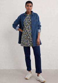 Dżinsowa koszula na kwiecistej tunice w zielono-białe wzory, zestawiona z ciemnoniebieskimi spodniami i białymi trampkami z teksturowaną podeszwą.