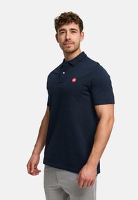 Puuvillainen laivastonsininen polopaita, lyhyet hihat, kaulus, vasemmassa rinnassa pieni punainen logo ja siisti, yksinkertainen muotoilu.