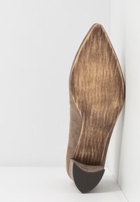 Chaussure en daim marron à bout pointu présentée debout, montrant une semelle en bois usée et un talon en caoutchouc noir sur fond blanc.