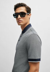 Grå polo med korta ärmar, marinblå krage och manschetter; bärs med stora svarta solglasögon i en elegant rektangulär form.
