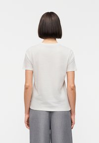 Personne aux cheveux courts, raides et foncés, portant un t-shirt blanc uni à manches courtes et un pantalon gris à jambes larges, vue de dos, sur un fond uni.
