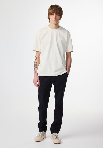 Creme kortærmet t-shirt parret med sorte bukser og lyse sneakers. Stoffet ser glat ud med en afslappet pasform og rund halsudskæring.